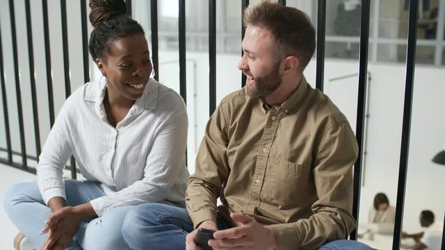Tracking shot of cheerful black woman and bearded man chatting while sitting on floor on mezzanine level of modern office or co-working space