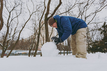 Smiling young man in blue winter warm clothes playing, making snowball, snowman in snowy park or forest outdoors. Winter fun, leisure on holidays. Love relationship family people lifestyle concept.