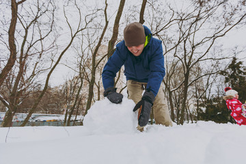 Cheerful young man in blue winter warm clothes playing, making snowball, snowman in snowy park or forest outdoors. Winter fun, leisure on holidays. Love relationship family people lifestyle concept.