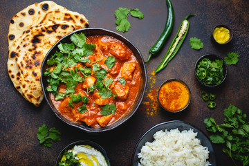 Traditional Indian dish Chicken tikka masala with spicy curry meat in bowl, basmati rice, bread naan, yoghurt raita sauce on rustic dark background, top view, close up. Indian style dinner from above - obrazy, fototapety, plakaty