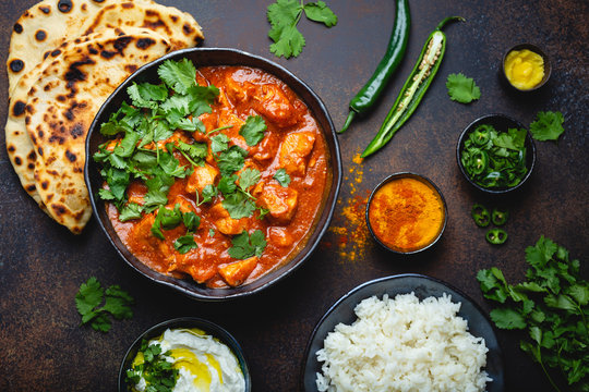 Traditional Indian dish Chicken tikka masala with spicy curry meat in bowl, basmati rice, bread naan, yoghurt raita sauce on rustic dark background, top view, close up. Indian style dinner from above
