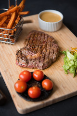 A platter of sirloin steak, salad, fries, sauce, tomato and mushrooms, served on a rustic wooden cutting board. Dark food photography with a mechanics workshop industrial setting.
