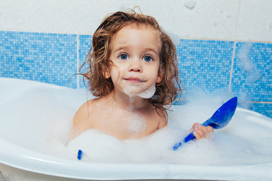 Fun cheerful happy toddler baby taking a bath playing with foam bubbles. Little child in a bathtub. Smiling kid in bathroom on blue background. Hygiene and health care.