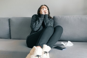 Young girl at home on the sofa in warm knitted sweater with handkerchief, sneezes. Flu and cold season