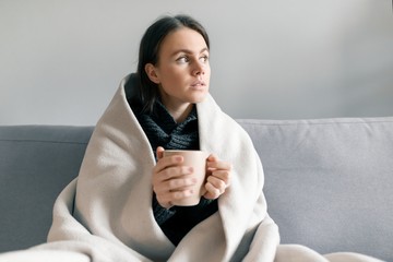 Autumn winter portrait of young girl resting at home on the sofa with cup of hot drink, under warm...
