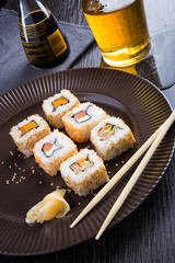 Sushi on a brown plate with a glass of beer, soy sauce and natural wooden chopsticks on a light wooden table.