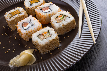Sushi on a brown plate with a glass of beer, soy sauce and natural wooden chopsticks on a light wooden table.