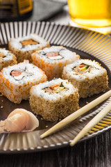 Sushi on a brown plate with a glass of beer, soy sauce and natural wooden chopsticks on a light wooden table.