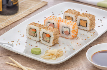 Sushi on a white square plate with soy sauce and natural wooden chopsticks on a light wooden table.