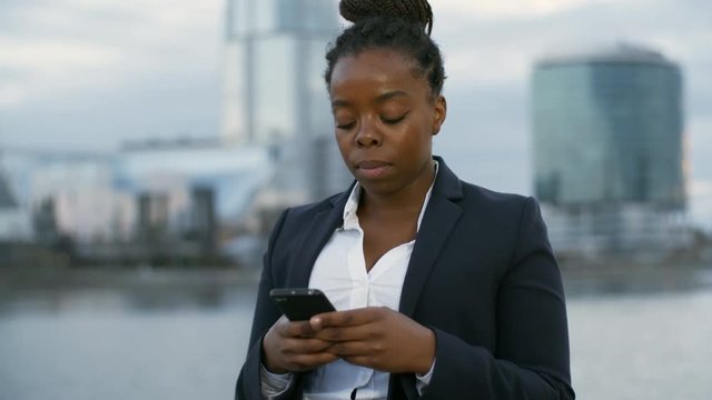 Panning of young African businesswoman standing near river and office buildings, holding phone and chatting with someone
