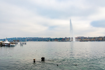 Outdoor tranquil scenery of THE JET D'EAU, famous water jet fountain on Geneva lake, embankment, boat and harbor along waterside with background of overcast sky.