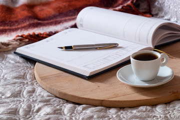 Cup with coffee, notepad, pen on the wooden desk on the bed.