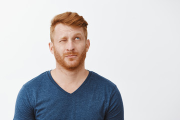 Fototapeta premium Hmm let us imagine. Portrait of dreamy cute adult redhead male with beard, looking up with one eye closed, lifting eyebrows, thinking or making up idea, picturing something over gray background