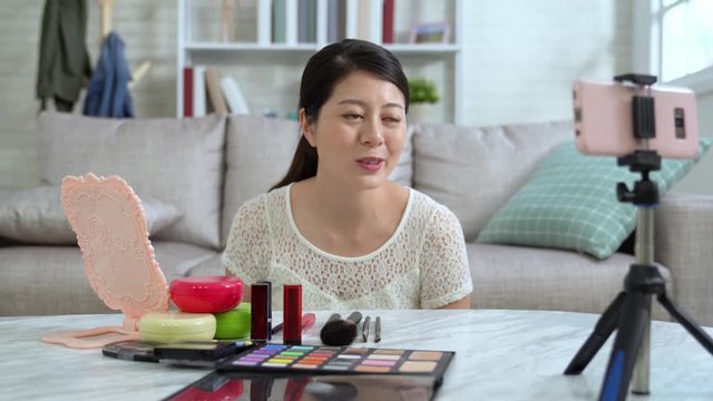 female blogger going to start teaching cosmetic makeup tutorial vlog online live video streaming. young girl waving hands to fans on internet looking at mobile phone in living room at home. 