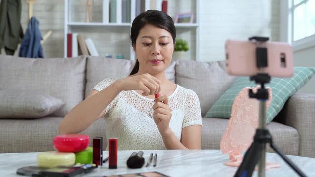 Young Asian blogger doing a cosmetic makeup tutorial vlog with brushes looking camera smartphone recording viral clip share on social media live video streaming Internet. girl showing lip gloss stain