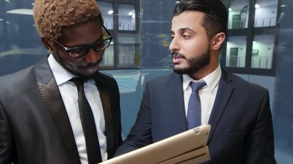Two multiethnic businessmen moving up in elevator with cup of coffee and tablet computer and discussing information about clients