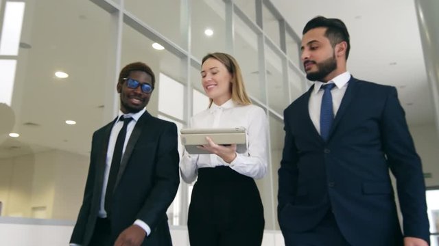 Track out shot of three mixed multiethnic business partners going inside modern futuristic building with tablet computer, smiling and speaking about work forecast