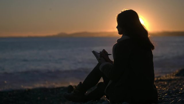 A pretty woman writing in her journal and drawing sketches on paper sitting on a scenic beach at sunset.