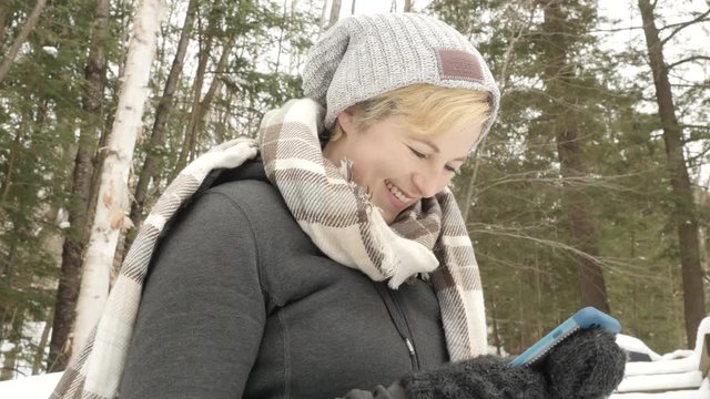 Low angle shot of blonde woman smiling as she types on her phone. Winter forest in background.