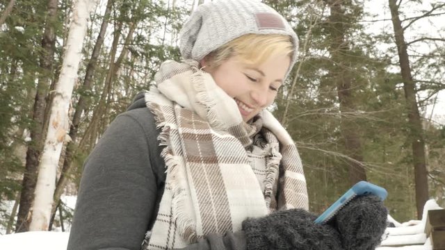 Low angle shot of blonde woman smiling as she texts on her phone. Winter forest in background.