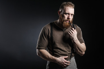 Lumberjack brutal bearded muscled man in brown shirt with smoking tube and suspenders on grey pants and axe on dark background