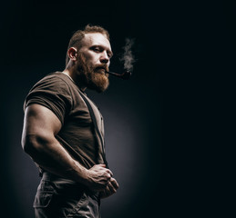 Lumberjack brutal red beard muscled man in brown shirt with smoking tube standing on dark background. Handsome man with red beard and moustache smoking pipe - obrazy, fototapety, plakaty