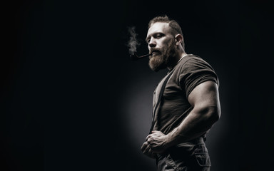 Lumberjack brutal red beard muscled man in brown shirt with smoking tube standing on dark background. Handsome man with red beard and moustache smoking pipe