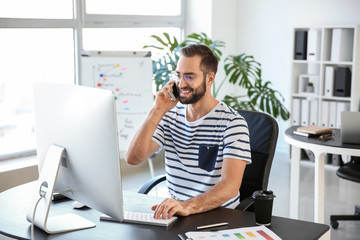 Young man talking on mobile phone while working on computer in office
