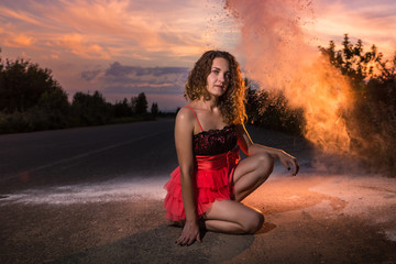 Fototapeta na wymiar Girl in red dress on the road and white flour cloud behind her in the evening at sunset