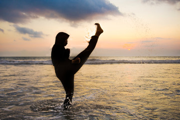 silhouette of young fit Muslim woman covered in Islam hijab head scarf training martial arts karate kick attack and fitness workout at beautiful beach sunset