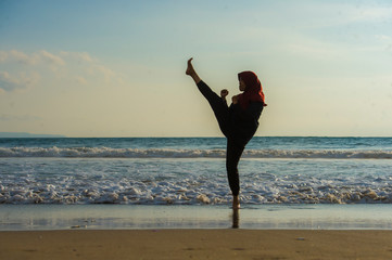 silhouette of young fit Muslim woman covered in Islam hijab head scarf training martial arts karate kick attack and fitness workout at beautiful beach sunset
