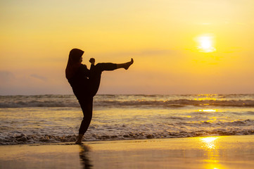 silhouette of young fit Muslim woman covered in Islam hijab head scarf training martial arts karate kick attack and fitness workout at beautiful beach sunset