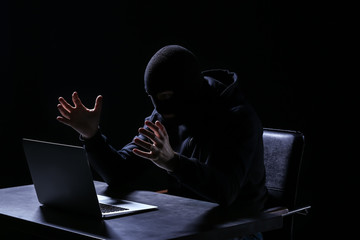 Emotional hacker with laptop sitting at table on dark background