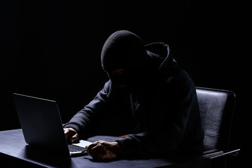 Professional hacker with laptop sitting at table on dark background