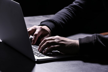 Professional hacker with laptop sitting at table on dark background, closeup