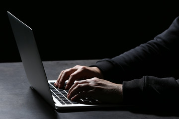 Professional hacker with laptop sitting at table on dark background, closeup