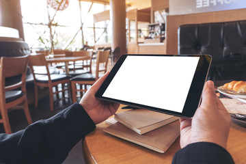 Mockup image of hands holding black tablet pc with blank screen with notebook and bread on wooden table in cafe