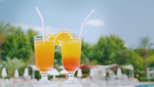 Two orange cocktails with straws and orange slices are on the beach table by the pool against the blue sky
