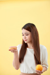 Portrait of a smiling young asian woman choosing between donut and orange isolated over yellow background