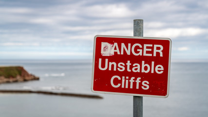 Sign: Danger unstable cliffs, with cliffs in the blurry background, seen in Berwick-upon-Tweed, Northumberland, England, UK