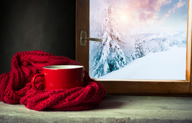 Fototapeta na wymiar Winter background - cup with candy cane, woolen scarf and gloves on windowsill and winter scene outdoors. Still life with concept of spending winter time at cozy home with cold weather outdoors