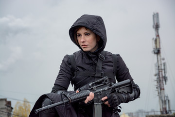 Young woman in modern black techwear style with rifle posing on the rooftop, portrait of redhead...