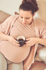 Pregnant woman drinking coffee while sitting on sofa at home.