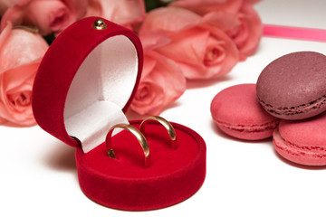 Box of wedding rings with pink and chocolate macarons on a background of bouquet of pink roses and pink ribbon. Macro view