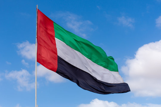 A United Arab Emirates (UAE Flag) blowing in the wind on a blue sky and puffy white cloud day.