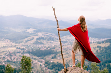 Unknown female in dress and red cape standing on stone at top of mountaing. Barefoot blonde woman...