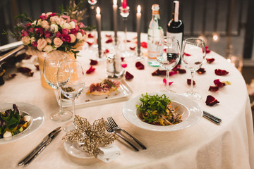 Romantic table setting with wine, beautiful flowers in box, empty glasses, rose petals and candles