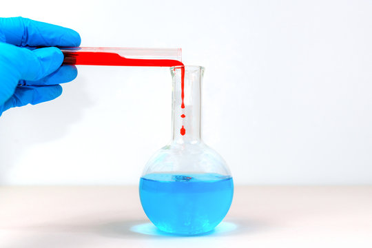 A hand wearing a blue glove pours red liquid from a test tube into a Florence flask with light blue liquid in it.