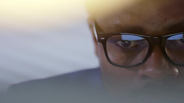 Close up tracking shot of eyes of black man in glasses sitting at desk in office and looking at computer screen