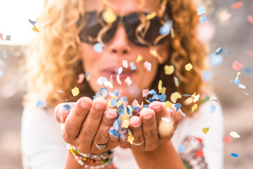 Close up of woman's habd and beautiful lady blowing out coloured carnival party confetti - focus on colors papers and hapiness and joyful lifestyle concept for happy people outdoor - obrazy, fototapety, plakaty
